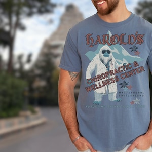 May include: A blue t-shirt with a graphic of a Yeti wearing a white coat and sunglasses. The text "Harold's Chiropractic & Wellness Center" is printed on the shirt. The text "Matterhorn, Switzerland" and "Adjusting spines since 1986" are also printed on the shirt.