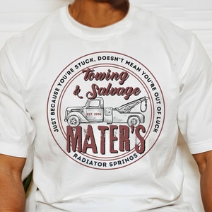 Puede incluir: Camiseta blanca con un gr&aacute;fico circular marr&oacute;n que presenta un cami&oacute;n de remolque vintage y el texto "Just because you're stuck, doesn't mean you're out of luck. Towing & Salvage Mater's Radiator Springs Est. 2006"