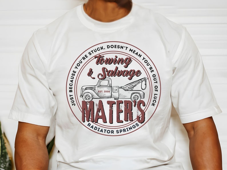 Puede incluir: Camiseta blanca con un gr&aacute;fico circular marr&oacute;n que presenta un cami&oacute;n de remolque vintage y el texto "Just because you're stuck, doesn't mean you're out of luck. Towing & Salvage Mater's Radiator Springs Est. 2006"