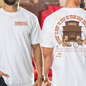 Könnte beinhalten: Weißes T-Shirt mit einem roten und schwarzen Grafikdruck eines Saloons mit dem Text "Pioneer Hall Since 1974" auf der Vorderseite und "Nothin' is goin' to stop us from hoop dee doo-in' tonight with Johnny Claire Jim Flora Dolly and Six Bits" auf der Rückseite.