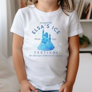 May include: White t-shirt with a blue graphic design featuring a castle, snowflakes, and the text "arendelle ELSA'S ICE Since 2013 FESTIVAL the cold never bothered me anyway".