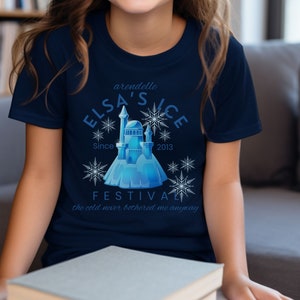May include: A navy blue t-shirt featuring a graphic of a blue ice castle and snowflakes. The text "Arendelle Elsa's Ice Festival Since 2013" and "The cold never bothered me anyway" is printed on the shirt.