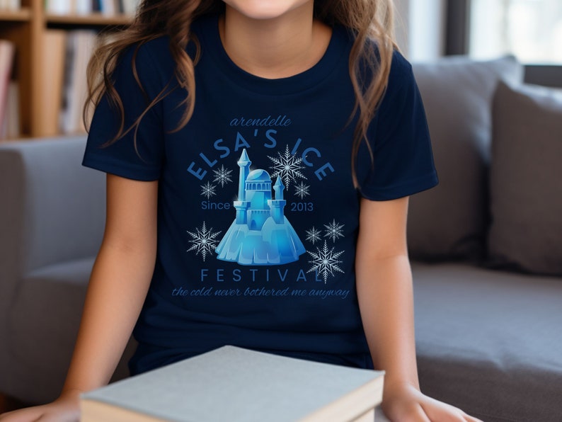 May include: A navy blue t-shirt featuring a graphic of a blue ice castle and snowflakes. The text "Arendelle Elsa's Ice Festival Since 2013" and "The cold never bothered me anyway" is printed on the shirt.