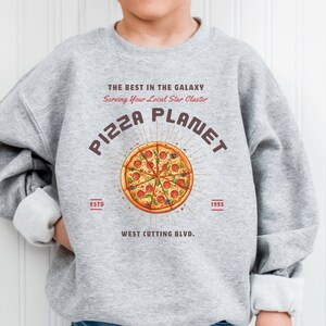 K&ouml;nnte beinhalten: Grauer Pullover mit einem Grafikdruck einer Pizza mit dem Text "PIZZA PLANET" in einem braunen Kreis mit Lichtstrahlen, die aus dem Kreis kommen. Der Text "THE BEST IN THE GALAXY" befindet sich &uuml;ber dem Pizzagrafi k und der Text "Serving Your Local Star Cluster" unter dem Pizzagrafi k. Der Text "ESTD" befindet sich links vom Pizzagrafi k und der Text "1995" rechts vom Pizzagrafi k. Der Text "WEST CUTTING BLVD." befindet sich unter dem Pizzagrafi k.