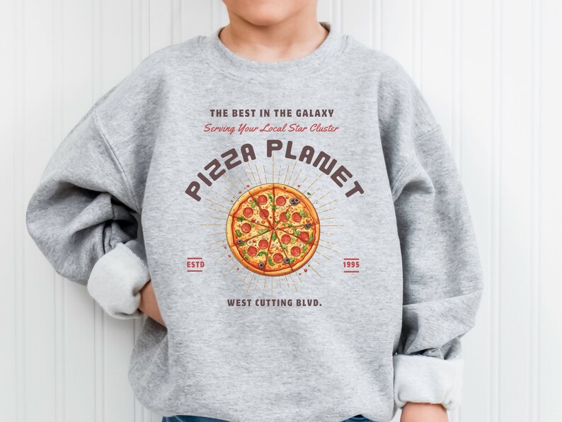 K&ouml;nnte beinhalten: Grauer Pullover mit einem Grafikdruck einer Pizza mit dem Text "PIZZA PLANET" in einem braunen Kreis mit Lichtstrahlen, die aus dem Kreis kommen. Der Text "THE BEST IN THE GALAXY" befindet sich &uuml;ber dem Pizzagrafi k und der Text "Serving Your Local Star Cluster" unter dem Pizzagrafi k. Der Text "ESTD" befindet sich links vom Pizzagrafi k und der Text "1995" rechts vom Pizzagrafi k. Der Text "WEST CUTTING BLVD." befindet sich unter dem Pizzagrafi k.