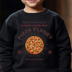 K&ouml;nnte beinhalten: Schwarzes Sweatshirt mit einem roten und wei&szlig;en Grafikdruck einer Pizza mit dem Text "Pizza Planet", "The Best in the Galaxy", "Serving Your Local Star Cluster", "Est. 1995" und "West Cutting Blvd."