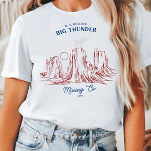 May include: White t-shirt featuring a desert landscape design with the text "B.T. Bullion Big Thunder Mining Co. 1979". The graphic is in shades of red and blue, depicting a mountain scene with a goat.