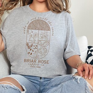 May include: A gray t-shirt with a brown graphic design featuring a circle with four quadrants. Each quadrant has a different design: a spinning wheel, a flower, a bird, and coffee beans. The text "Wake up and smell the roses" is written around the circle. Below the circle is the text "Briar Rose coffee co. since 1959."