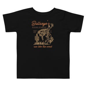 May include: Black toddler t-shirt with a tan graphic of a cowboy riding a bucking bronco. The shirt features the text "Bullseye's Riding Stables" and "run like the wind". A cactus is also included in the design.