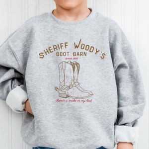 Puede incluir: Sudadera gris con el texto "SHERIFF WOODY'S BOOT BARN since 1995" en letras marrones. Debajo del texto hay una ilustraci&oacute;n marr&oacute;n de una bota de vaquero. La frase "There's a snake in my boot" est&aacute; escrita debajo.