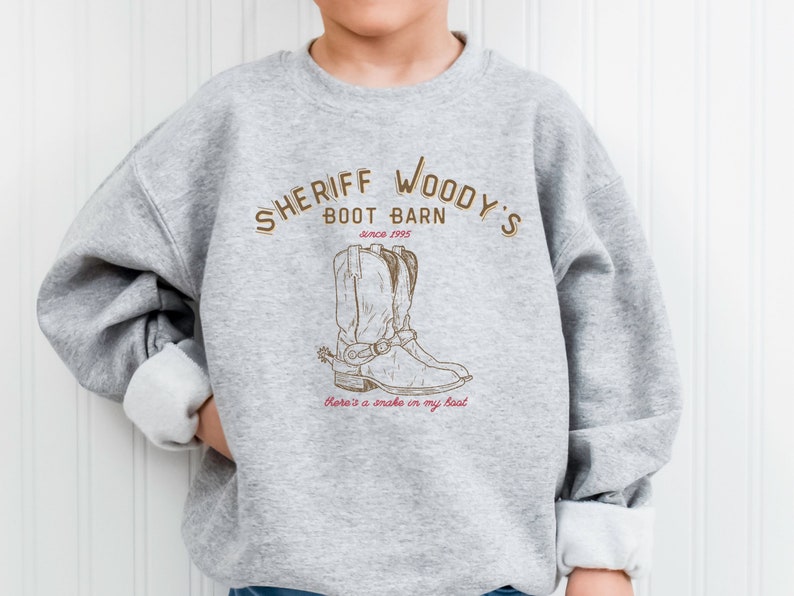 Puede incluir: Sudadera gris con el texto "SHERIFF WOODY'S BOOT BARN since 1995" en letras marrones. Debajo del texto hay una ilustraci&oacute;n marr&oacute;n de una bota de vaquero. La frase "There's a snake in my boot" est&aacute; escrita debajo.