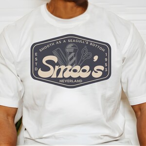 May include: White t-shirt with a navy blue logo that says "Smooth as a Seagull's Bottom" and "Smee's" with a vintage style font. The logo also includes a barber pole, comb, and scissors. The text "Est. 1953" and "Neverland" are also included in the logo.