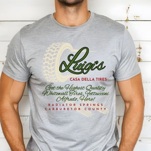 May include: A grey t-shirt with a green and white logo that says "Luigi's Casa Della Tires". The logo features a tyre with a tread pattern. The text below the logo says "Get the Highest Quality Whitewall Tyres, Fettuccini Alfredo, Here! Radiator Springs Carburetor County."