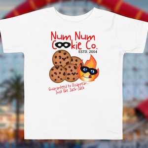 May include: White t-shirt with a cartoon design of chocolate chip cookies and a flame character wearing a mask. The shirt has the text "Num Num Cookie Co. ESTD. 2004" and "Guaranteed to Disappear - Just like Jack-Jack."
