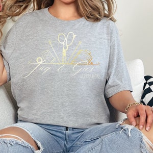 May include: A gray t-shirt with a gold design of sewing tools and the text "Jay & Girs Alterations".
