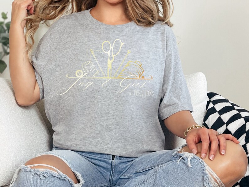 May include: A gray t-shirt with a gold design of sewing tools and the text "Jay & Girs Alterations".
