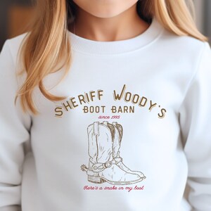 Puede incluir: Sudadera blanca con el texto "SHERIFF WOODY'S BOOT BARN since 1995" en letras marrones. Debajo del texto hay una ilustraci&oacute;n marr&oacute;n de una bota de vaquero con una espuela. La frase "there's a snake in my boot" est&aacute; escrita en rojo.