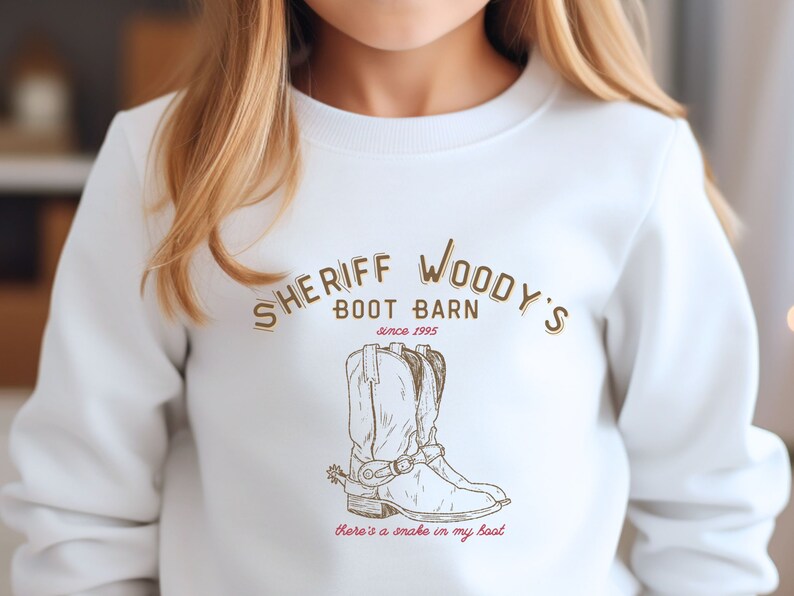 Puede incluir: Sudadera blanca con el texto "SHERIFF WOODY'S BOOT BARN since 1995" en letras marrones. Debajo del texto hay una ilustraci&oacute;n marr&oacute;n de una bota de vaquero con una espuela. La frase "there's a snake in my boot" est&aacute; escrita en rojo.