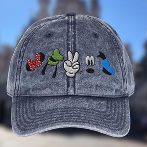 May include: A denim blue baseball cap with embroidered cartoon character elements. The design includes a red and black polka dot bow, a green glove, a white peace sign, and a blue and black element.