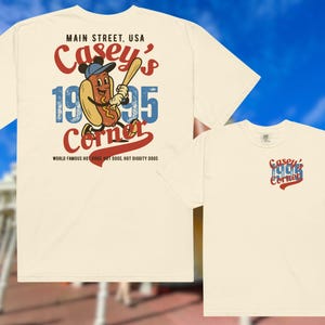 May include: Cream-colored t-shirt featuring a cartoon hot dog character in a baseball cap holding a bat. The shirt displays "Casey's Corner" with "1995" and "Main Street, USA" text. The back of the shirt has the same design.