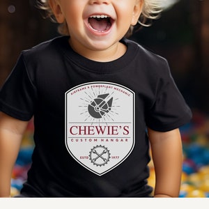 Puede incluir: Una camiseta negra con un diseño gráfico blanco y gris. El gráfico presenta un logotipo de estilo vintage con el texto "Chewie's Custom Hangar" y una ilustración de una nave espacial. El texto "Airframe & Powerplant Mechanic" está por encima del logotipo, y "Estd 1977" está por debajo del logotipo.