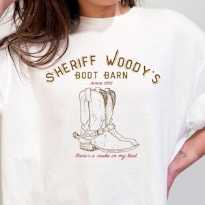May include: White t-shirt with a brown graphic of a cowboy boot with the text "Sheriff Woody's Boot Barn since 1995" and "There's a snake in my boot".