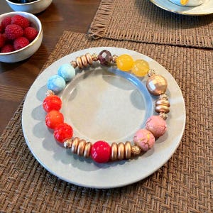 May include: A colorful beaded bracelet with a variety of round and textured beads in shades of red, orange, yellow, blue, and pink. The bracelet also features gold-colored metal accents. The bracelet is displayed on a light gray plate.