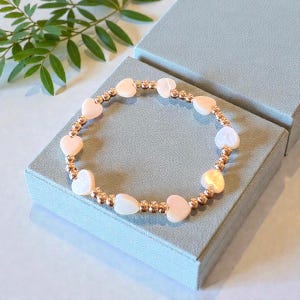 May include: A delicate bracelet featuring heart-shaped beads in soft pink and white, interspersed with small, gold-colored beads. The bracelet is displayed on a light blue box, with green foliage in the background.