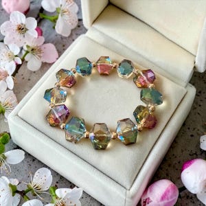May include: A circular bracelet composed of faceted, iridescent glass beads that shift between shades of green, gold, and purple. Small, round, metallic gold-toned spacer beads are positioned between each larger, multi-faceted crystal bead.