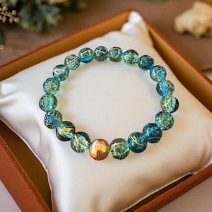 May include: A beaded bracelet featuring translucent teal and gold-veined beads. The bracelet includes a few larger, gold-colored beads. The bracelet is displayed on a white satin pillow inside a brown box.