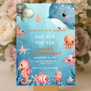Può includere: Biglietto d'invito blu a tema sottomarino. Presenta illustrazioni di creature marine come una balena, una tartaruga, un cavalluccio marino e un polpo. Il testo recita "ONE-DER THE SEA ANNA'S 1ST BIRTHDAY" con i dettagli dell'evento.