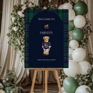 May include: A navy blue and green plaid welcome sign on a wooden easel. The sign reads "Welcome to Fareed's First Birthday Party" and features a teddy bear illustration. The background includes white and green balloons and floral arrangements.