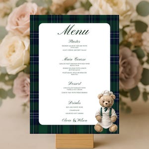 May include: A wedding menu card with a green and navy plaid border. The menu includes starters, main courses, desserts, and drinks. A teddy bear wearing a chef's hat and apron is on the right. The names "Olivia & Wilson" are at the bottom.