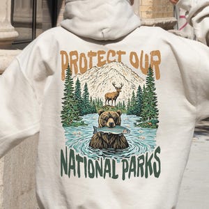 May include: A cream-coloured hoodie with a graphic print of a bear fishing in a lake with mountains and trees in the background. The text "Protect Our National Parks" is printed below the image.
