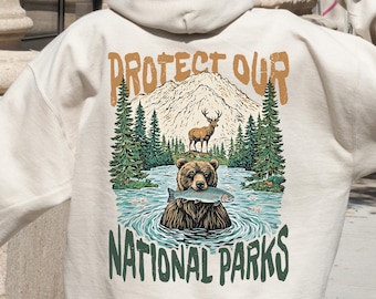 Puede incluir: Una sudadera con capucha de color crema con un estampado gráfico de un oso pescando en un lago con montañas y árboles en el fondo. El texto "Protect Our National Parks" está impreso debajo de la imagen.