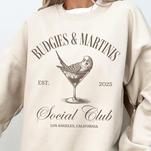 May include: A beige sweatshirt with the text "Budgies & Martinis Social Club Est. 2025 Los Angeles, California" and a graphic of a budgie in a martini glass.
