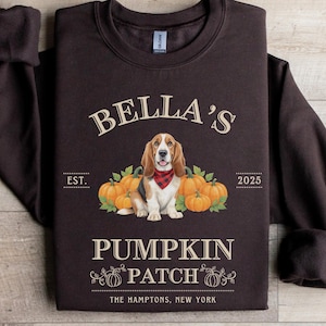 Könnte beinhalten: Ein dunkelbraunes Sweatshirt mit dem Text "Bella's Pumpkin Patch" und einer Grafik eines Basset Hounds mit rotem Halstuch, umgeben von Kürbissen. Der Text "The Hamptons, New York" befindet sich unten.