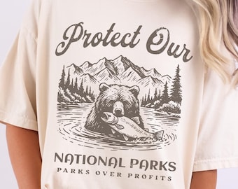 Könnte beinhalten: Ein beiges T-Shirt mit einem braunen Bären, der einen Fisch hält. Der Text "Protect Our National Parks" und "Parks Over Profits" ist auf dem T-Shirt aufgedruckt. Das T-Shirt ist mit "Ivory" gekennzeichnet.