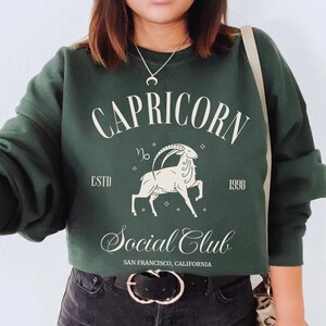 May include: A dark green sweatshirt with a white graphic print of a Capricorn zodiac sign and the text "Capricorn Social Club Est. 1998 San Francisco, California".