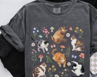 May include: A dark gray t-shirt with a graphic print of cats and flowers. The cats are in various poses, including sitting, lying down, and standing. The flowers are in shades of pink, purple, and yellow.