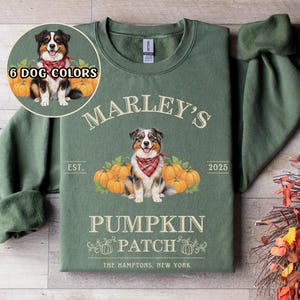 Peut inclure: Sweat-shirt vert sauge avec l'inscription "Marley's Pumpkin Patch" et un motif de chien. Le design comprend des citrouilles et le texte "The Hamptons, New York". Un petit cercle affiche "6 Dog Colors".