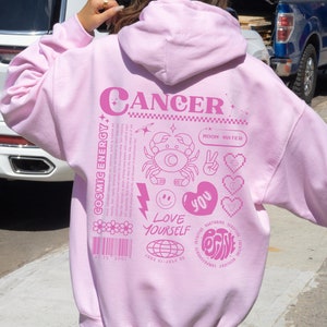 May include: Pink hoodie with a graphic design featuring the zodiac sign Cancer, the text "Cancer", "Moon Water", "Cosmic Energy", "Love Yourself", "You", and "Positive".