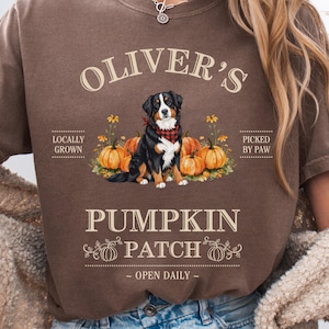 May include: Brown t-shirt with the text "Oliver's Pumpkin Patch" and a graphic of a Bernese Mountain Dog wearing a red bandana, surrounded by pumpkins. The shirt also includes the phrases "Locally Grown" and "Picked by Paw."