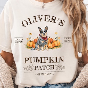 May include: Cream-colored t-shirt featuring a dog illustration with pumpkins and text. The text reads "Oliver's Pumpkin Patch" with additional text "Locally Grown", "Picked by Paw", and "Open Daily". The dog wears a red bandana.