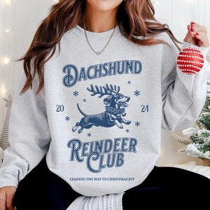 May include: A grey sweatshirt with a blue graphic of a dachshund dressed as a reindeer. The text on the sweatshirt reads "Dachshund Reindeer Club" and "Leading the way to Christmas joy".