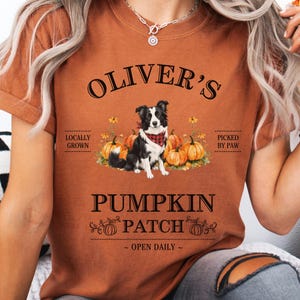 May include: A burnt orange t-shirt with the text "Oliver's Pumpkin Patch" and a graphic of a black and white dog wearing a red bandana, surrounded by pumpkins. The shirt also includes the phrases "Locally Grown" and "Picked by Paw".