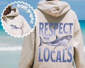 May include: Beige hoodie with a shark graphic. The back of the hoodie features a large shark illustration with the text "RESPECT THE LOCALS" in blue. A smaller shark graphic is on the front.