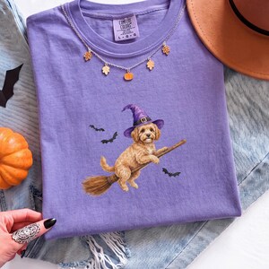 Peut inclure: Un t-shirt à col rond violet avec un motif représentant un chien doré portant un chapeau de sorcière violet et chevauchant un balai en bois, accompagné de trois petites chauves-souris noires. L'étiquette Comfort Colors est visible au col.