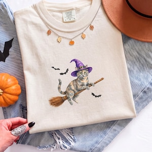 Pode incluir: Uma t-shirt de cor creme com o gráfico de um gato a usar um chapéu de bruxa roxo e a montar uma vassoura, rodeado por pequenos morcegos pretos. A t-shirt tem um colar prateado com pequenos pingentes de folhas e abóboras no colarinho.