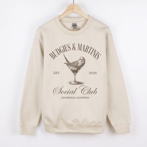 May include: A beige sweatshirt with a brown graphic print of a budgie in a martini glass. The text on the graphic reads "Budgies & Martinis Social Club Est. 2025 Los Angeles, California."